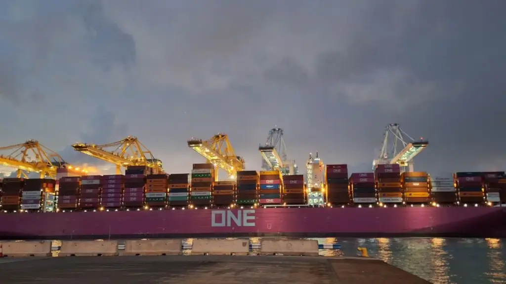 A imagem retrata um navio porta-contêineres de grande porte atracado em um terminal portuário durante a noite, com centenas de contêineres organizados e guindastes operando de forma sincronizada, evidenciando a complexidade e a continuidade da logística internacional, onde cada etapa — do planejamento ao embarque, do controle documental à execução operacional — precisa estar integrada para garantir visibilidade, previsibilidade e eficiência em uma operação logística verdadeiramente end-to-end.