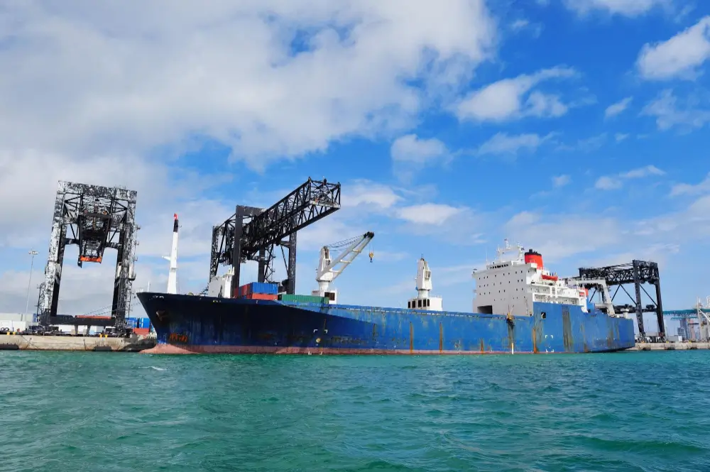 Navio cargueiro azul atracado em terminal portuário com guindastes de carga ao fundo, representando o processo de importação sujeito à fiscalização aduaneira e aos canais de conferência da Receita Federal no Brasil.